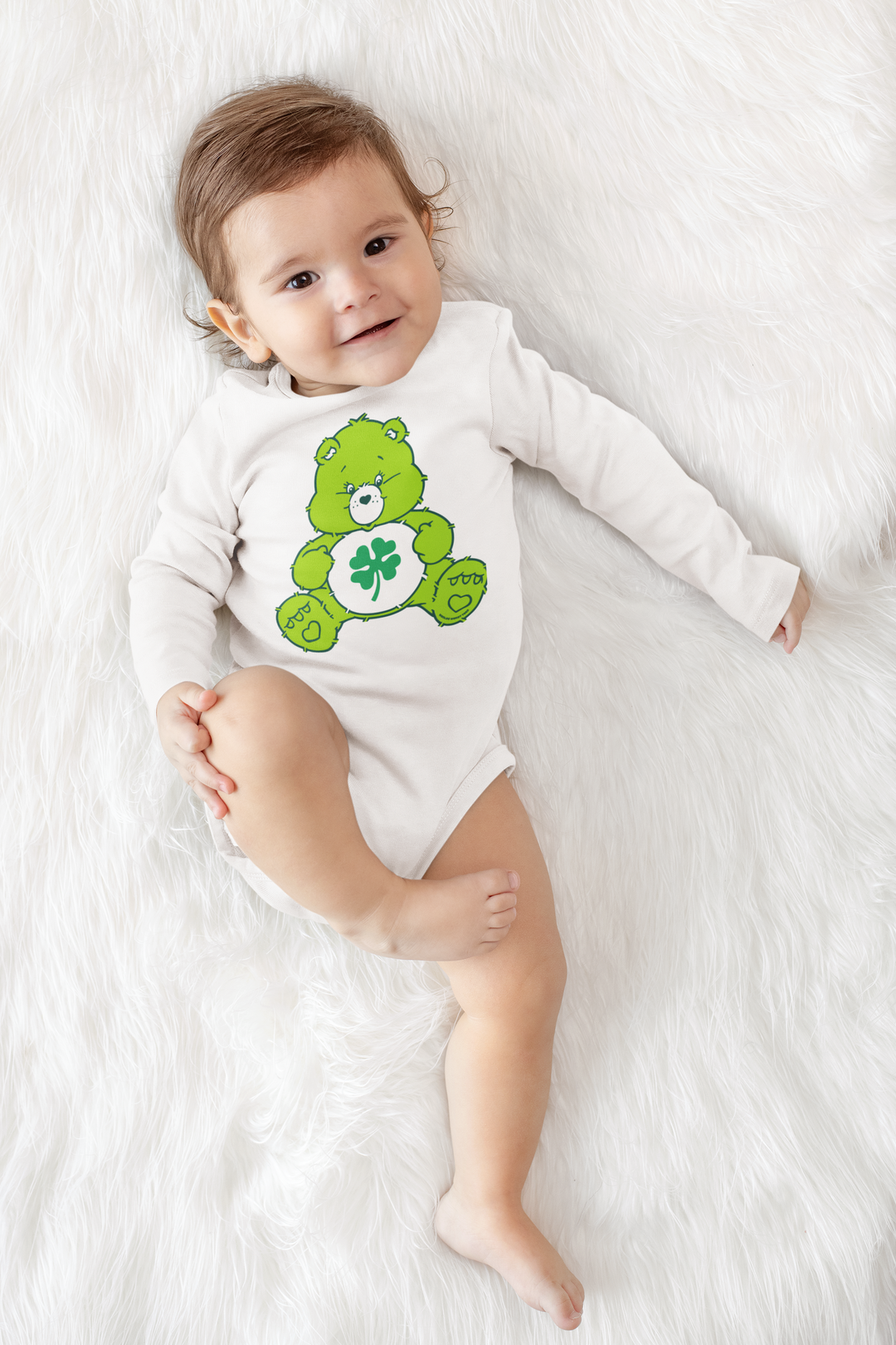 A Lucky Bear Onesie for infants, featuring a baby lying on a white blanket with a green teddy bear and clover. Made of soft cotton with plastic snaps for easy changing. From Worlds Worst Tees.