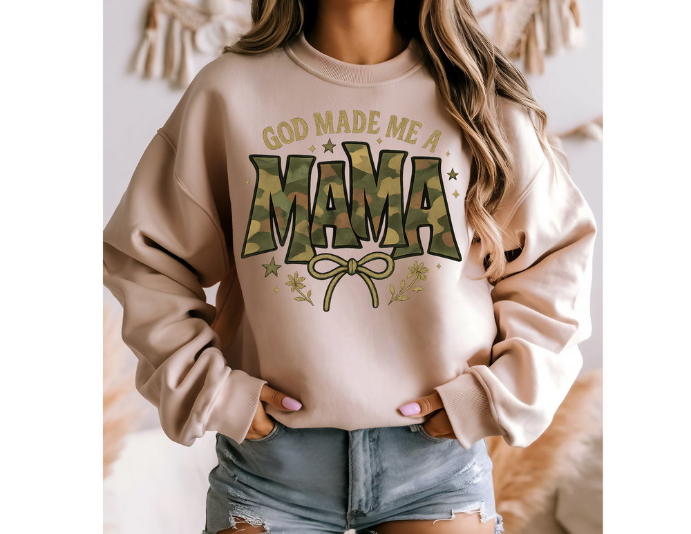 Person wearing God Made Me a Mama crew sweatshirt with camouflage MAMA text, star and floral accents, paired with distressed denim shorts. Neutral home décor in background.