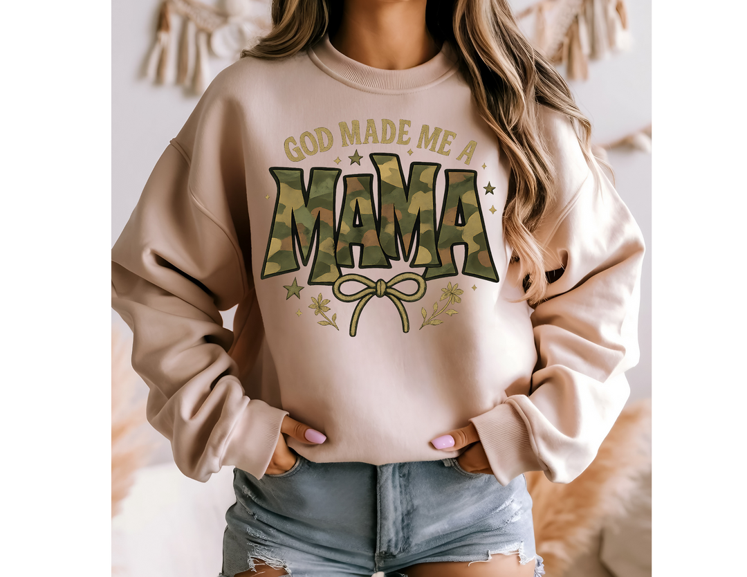Person wearing God Made Me a Mama crew sweatshirt with camouflage MAMA text, star and floral accents, paired with distressed denim shorts. Neutral home décor in background.