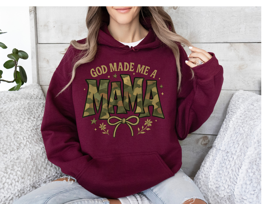 Person wearing God Made Me a Mama hoodie with camouflage MAMA text, bow, stars, and floral accents, paired with distressed jeans, sitting casually on a cozy couch.