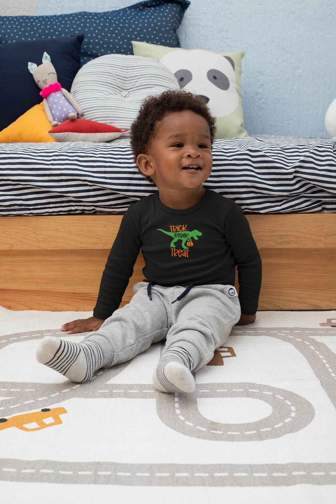A Trick Rawr Treat Long Sleeve Onesie featuring a child with a green dinosaur, lying on a bed, and sitting on the floor. Made of 100% combed ring-spun cotton for durability and comfort. Plastic snaps for easy changing.
