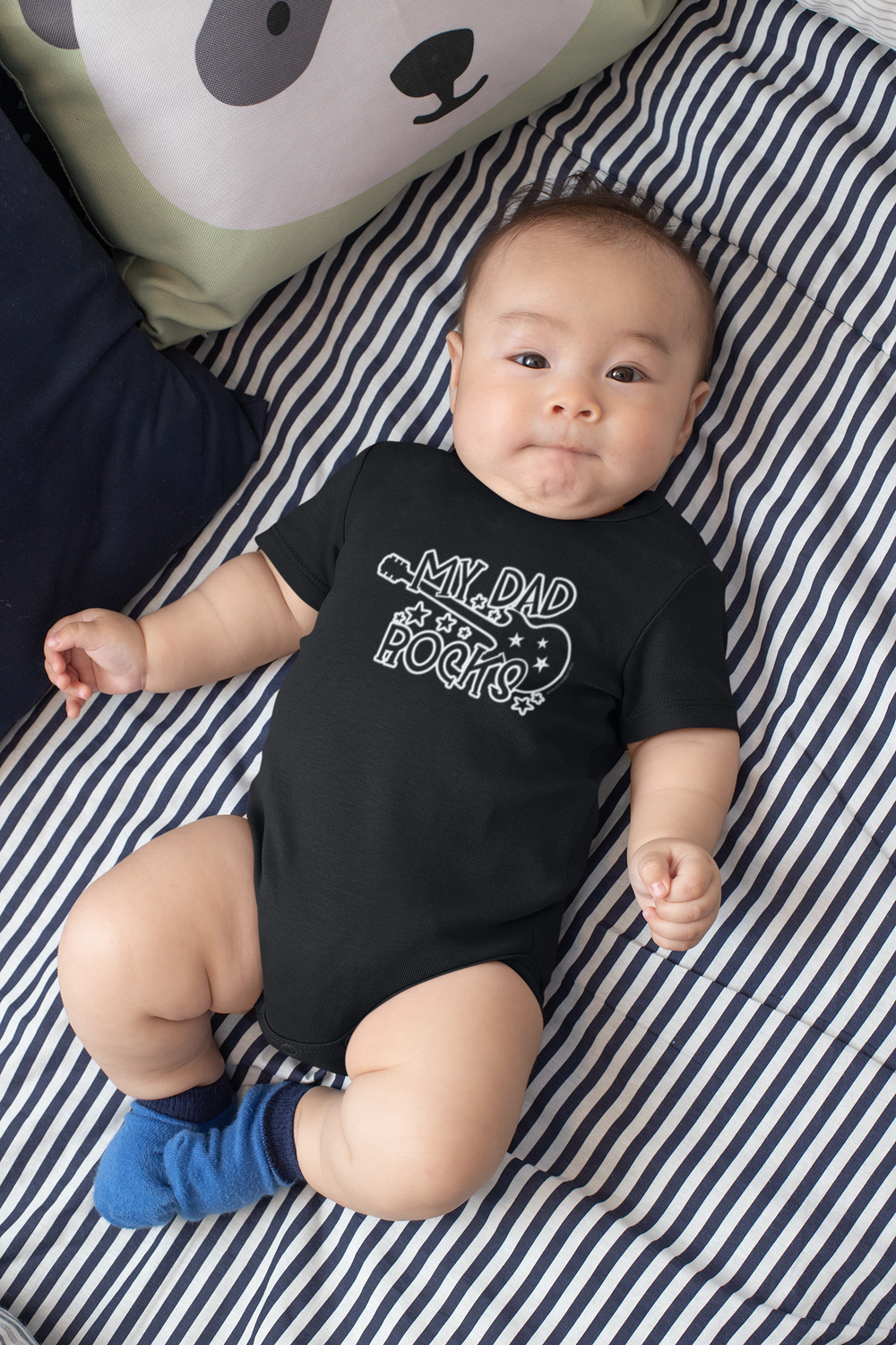 A baby lies on a striped blanket, wearing a My Dad Rocks Onesie from Worlds Worst Tees. This infant fine jersey bodysuit is 100% cotton, featuring ribbed bindings and plastic snaps for easy changes.