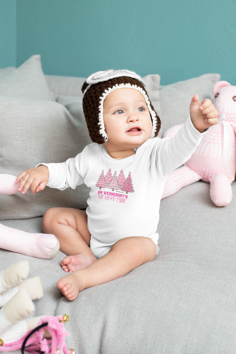 A baby wearing a knit hat sitting on a couch, pointing at something. Infant long sleeve bodysuit in pink, featuring plastic snaps for easy changing. Durable and soft 100% cotton fabric.