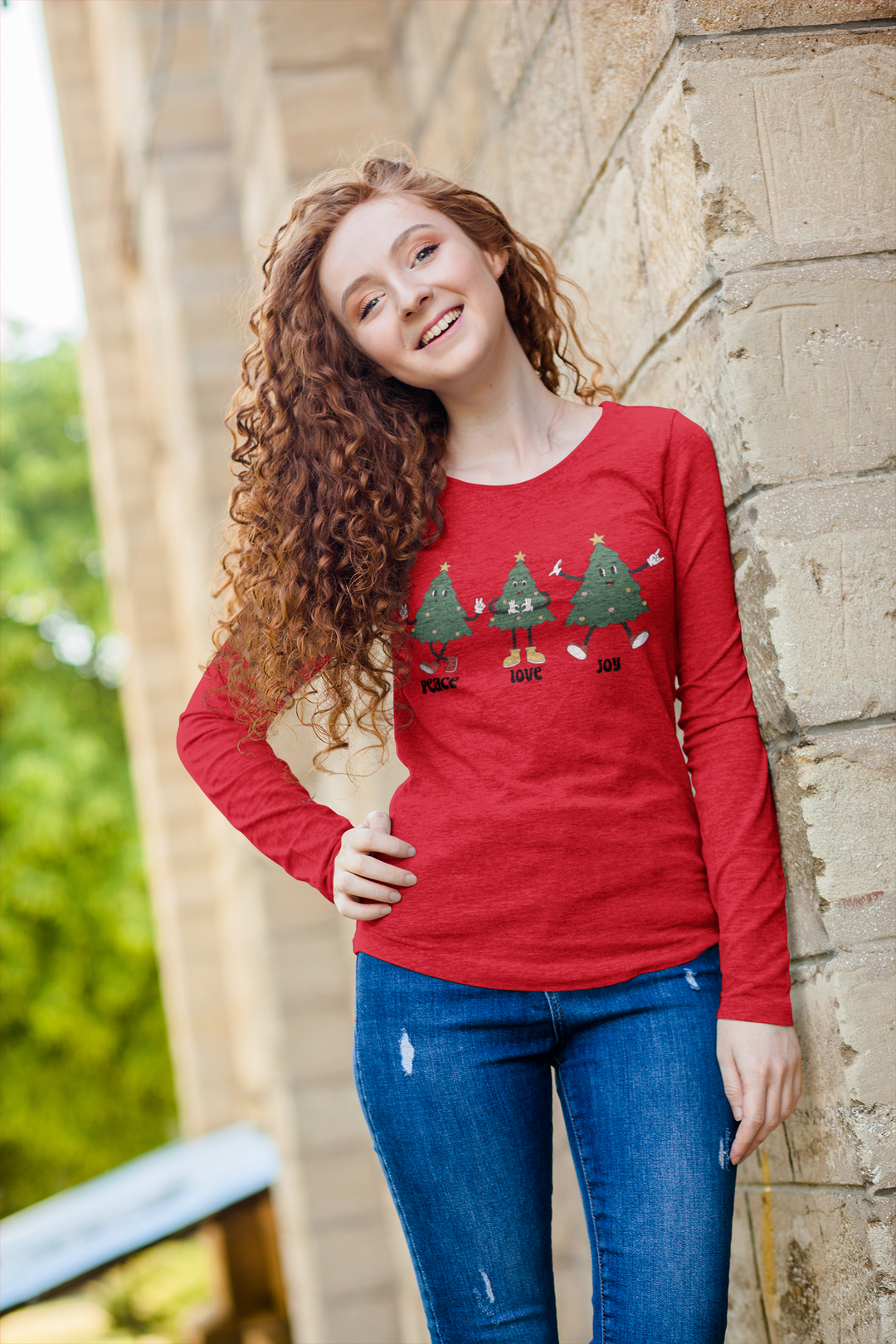 A woman in a red Peace Love Joy Long Sleeve Tee, featuring long curly hair, standing casually next to a brick wall. Classic fit, no side seams, 100% cotton for comfort. From Worlds Worst Tees.