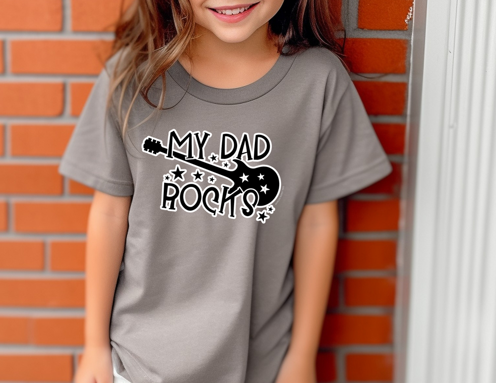 A girl in a casual dress stands in front of a brick wall. My Dad Rocks Kids Tee: 100% combed cotton, light fabric, classic fit, soft-washed, and garment-dyed for comfort and agility.