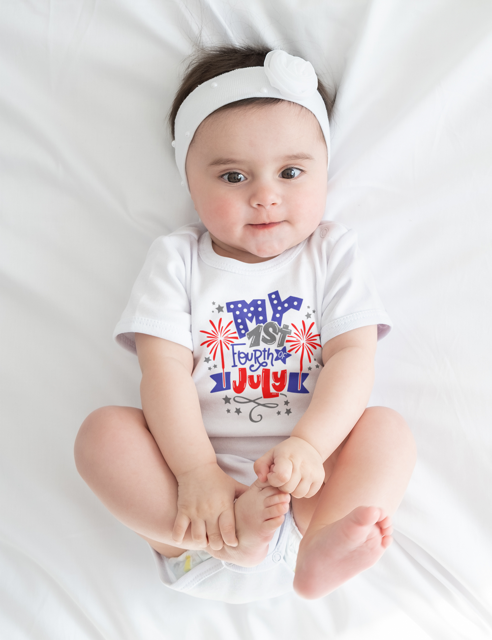 A baby lies on a white sheet, wearing a My 1st 4th of July Onesie. This infant fine jersey bodysuit is 100% cotton, featuring ribbed knit bindings and plastic snaps for easy changes. Durable and soft for little ones.