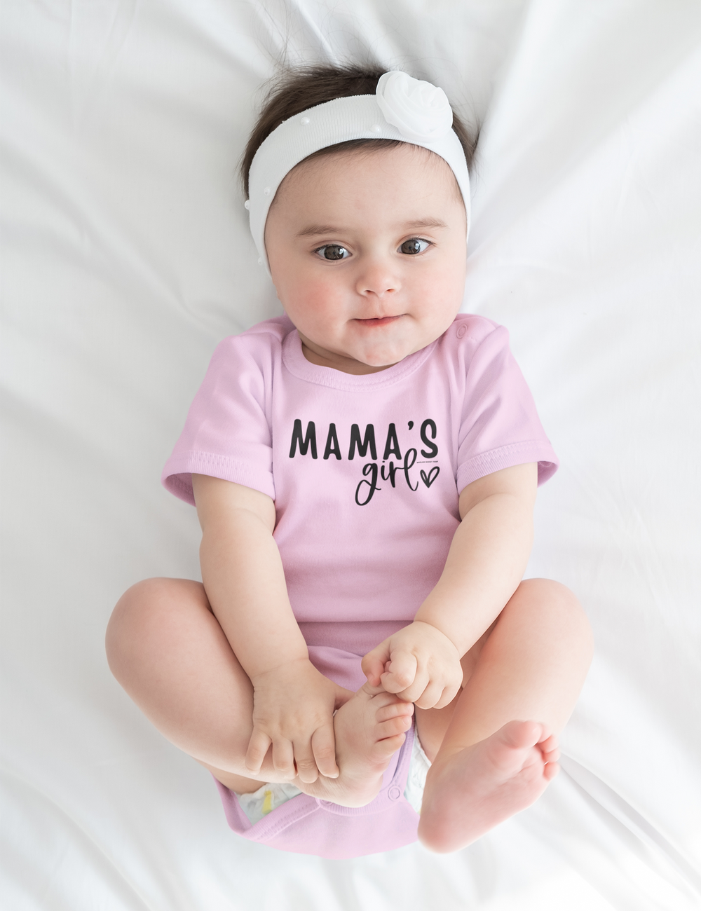 Infant fine jersey bodysuit featuring Mama's Girl Onesie. 100% cotton fabric, ribbed knit bindings, plastic snaps for easy changing. Close-up of a baby in a pink shirt, with a focus on the baby's face, feet, and hands.
