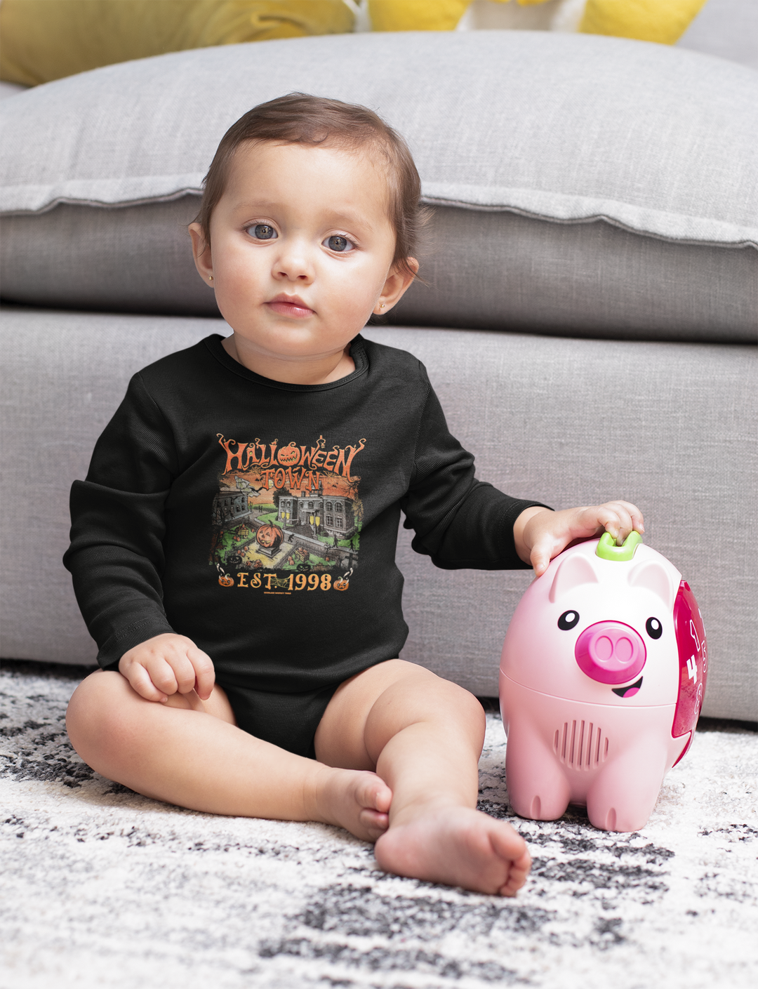 A baby in a black shirt with a house and pumpkins drawing, sitting with a toy pig. Halloweentown Long Sleeved Onesie for infants, featuring ribbed bindings and plastic snaps for easy changing.