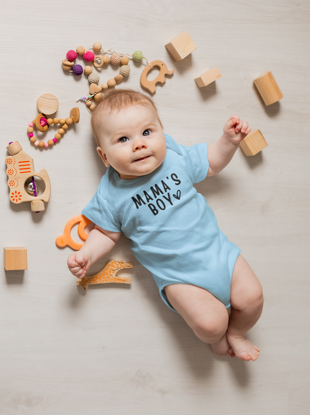 A baby lies on its back surrounded by toys, wearing a Mama's Boy Onesie. This infant fine jersey bodysuit is 100% cotton, featuring ribbed knitting for durability and plastic snaps for easy changing access. Dimensions vary by size.