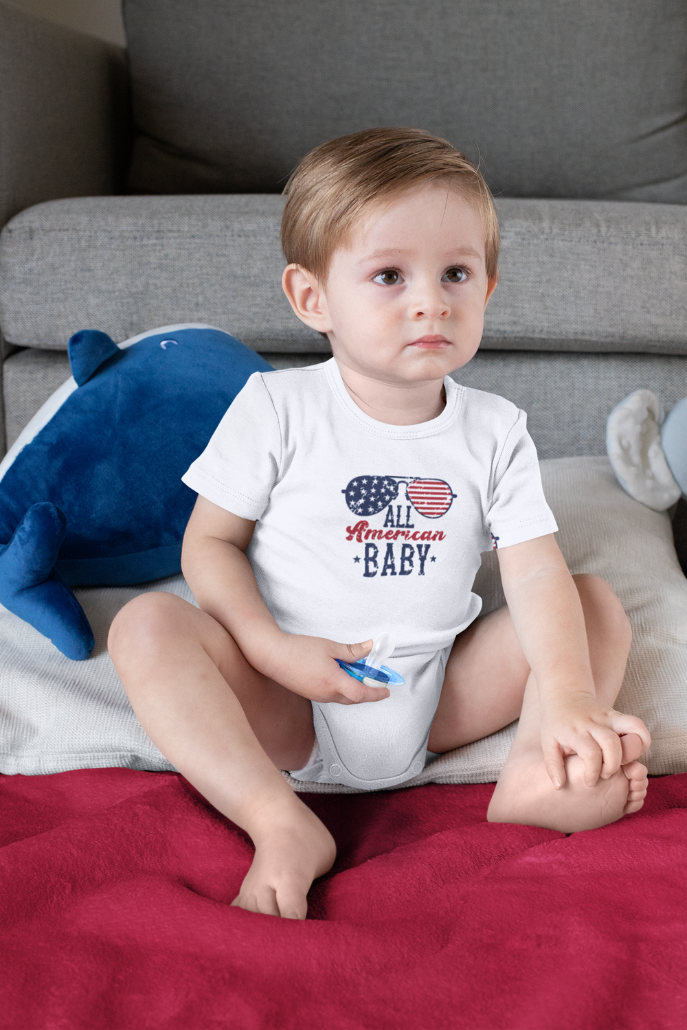 A baby in a white shirt sits comfortably on a couch, showcasing the All American Baby Onesie. Durable and soft, this infant fine jersey bodysuit features ribbed knitting for durability and plastic snaps for easy changing access.