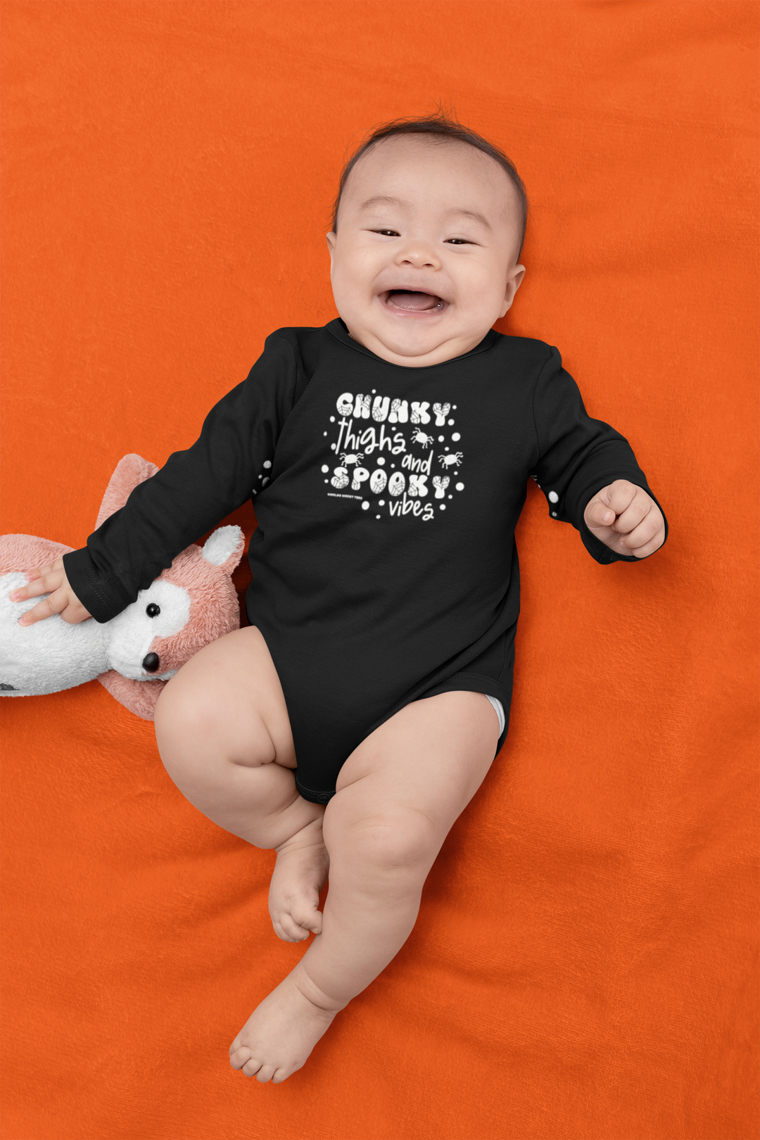 A baby lies on a blanket with a stuffed animal, showcasing the Chunky Thighs and Spooky Vibes Long Sleeved Onesie from Worlds Worst Tees. This infant bodysuit in 100% cotton features plastic snaps for easy changing.