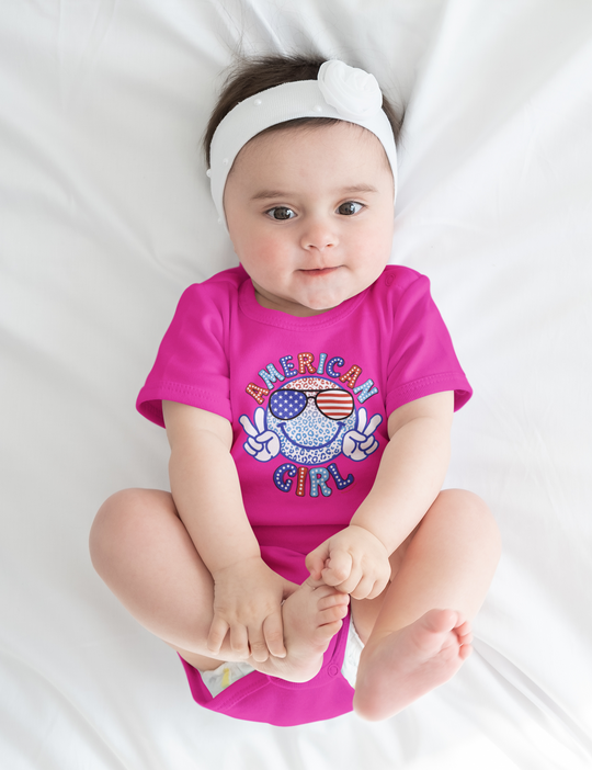 A baby lies on a white bodysuit with a Peace American Girl design. 100% cotton fabric, ribbed knit bindings, and plastic snaps for easy changing access. From Worlds Worst Tees.