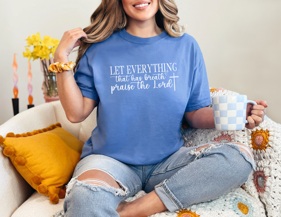 A woman in a cozy setting, holding a cup, showcases the Let Everything That Has Breath Praise the Lord Tee. This garment-dyed, ring-spun cotton t-shirt offers a relaxed fit and durable double-needle stitching.
