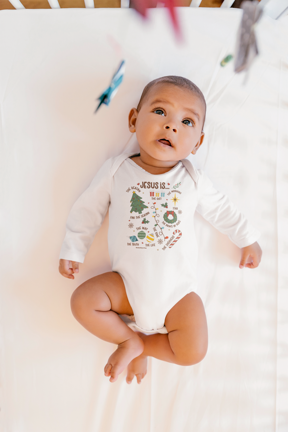 A baby lies on a white sheet, wearing a Jesus is Christmas onesie. This infant long sleeve bodysuit is soft, durable, and easy to change with plastic snaps. Made of 100% combed ring-spun cotton.