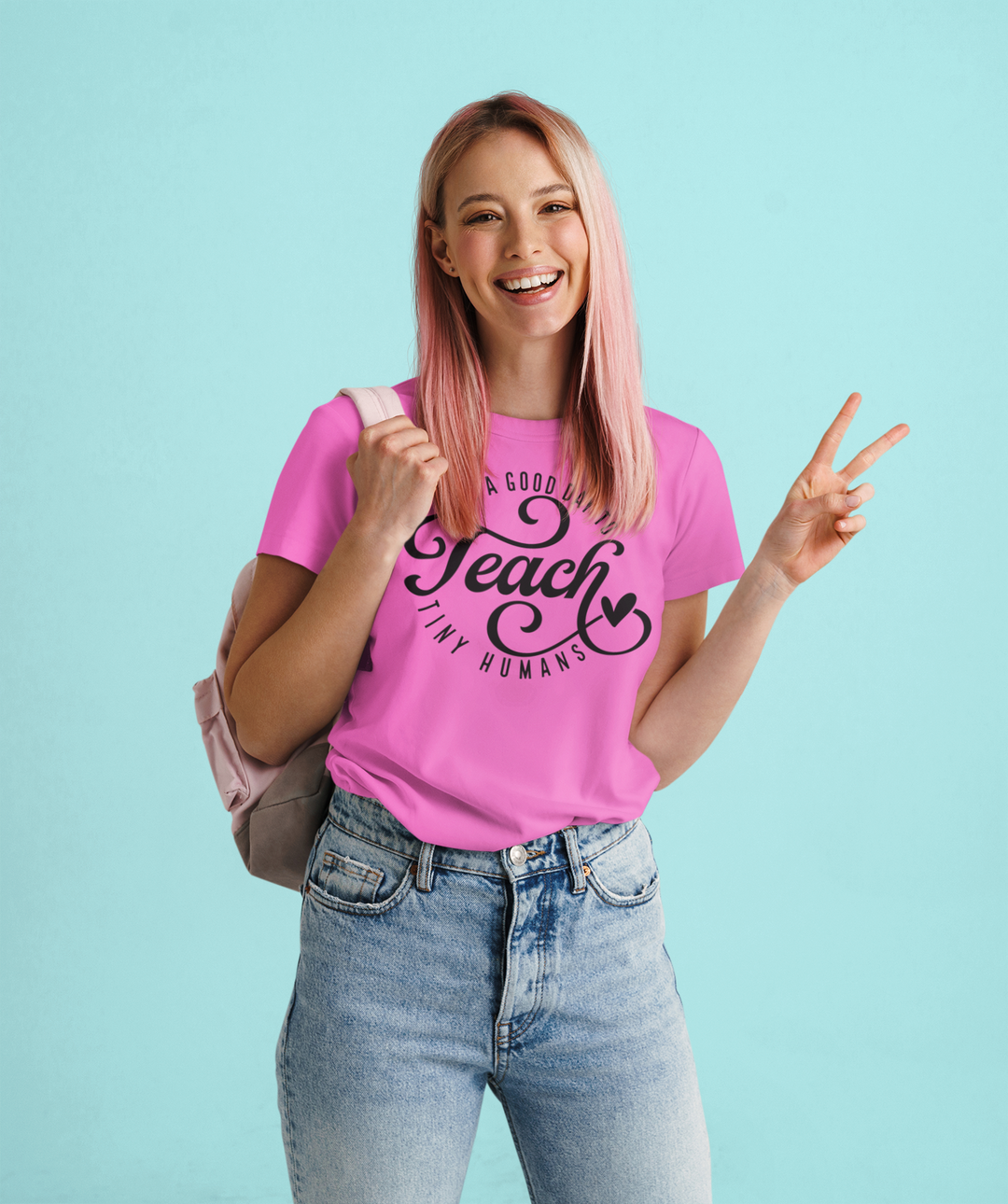 A smiling woman in a pink shirt holds up two fingers, showcasing the Teach Tiny Humans Tee. Unisex sweatshirt made of 80% ring-spun cotton, 20% polyester, with a relaxed fit and rolled-forward shoulder.