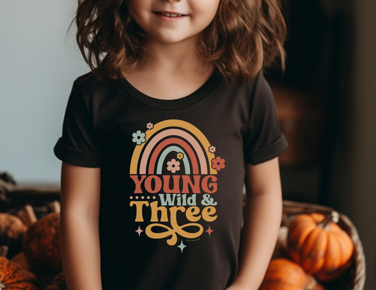 Young Wild and Three Toddler Tee featuring a girl standing by pumpkins, a child in a black shirt, and a rainbow on black. Soft 100% cotton, light fabric, tear-away label, perfect for sensitive skin.
