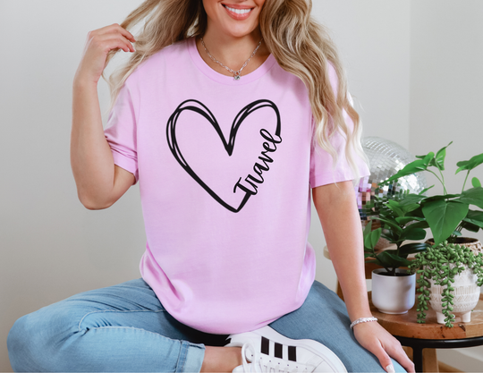 Woman in light-blue jeans wearing a pink Travel Tee with heart design, sitting cross-legged in a cozy interior with potted plants and a disco ball.