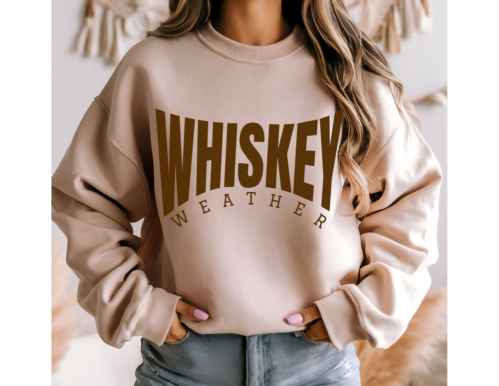 Person wearing a light beige Whiskey Weather Crew sweatshirt with brown lettering, paired with light blue jeans, showcasing casual, cozy style.
