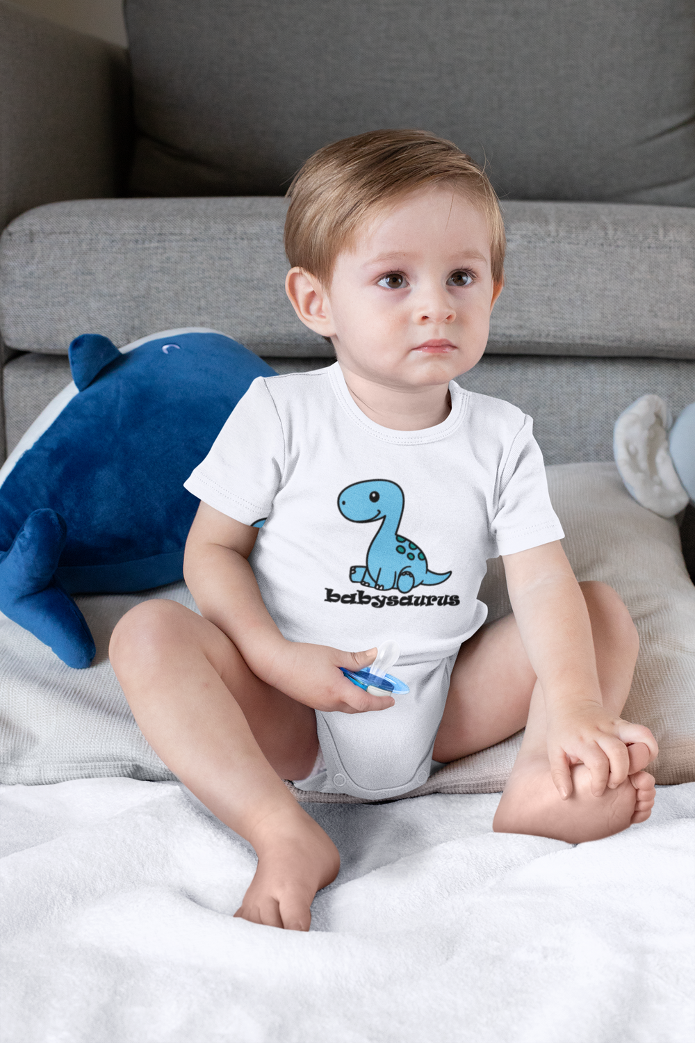 A baby wearing a Babysaurus Onesie, sitting on a bed, with a blue dinosaur graphic. Infant fine jersey bodysuit, 100% cotton, ribbed knitting for durability, plastic snaps for easy changing access. From Worlds Worst Tees.