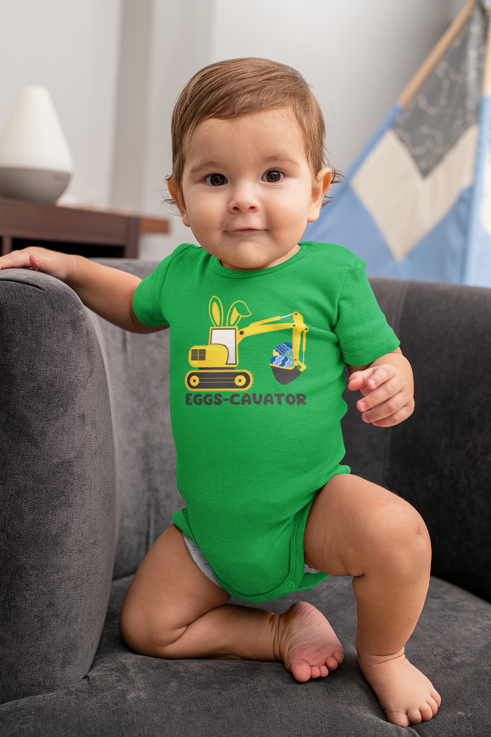 A baby in a green shirt wearing an Eggs-Cavator Onesie. Infant fine jersey bodysuit made of 100% cotton (fiber content may vary), with ribbed knitting for durability and plastic snaps for easy changing access.