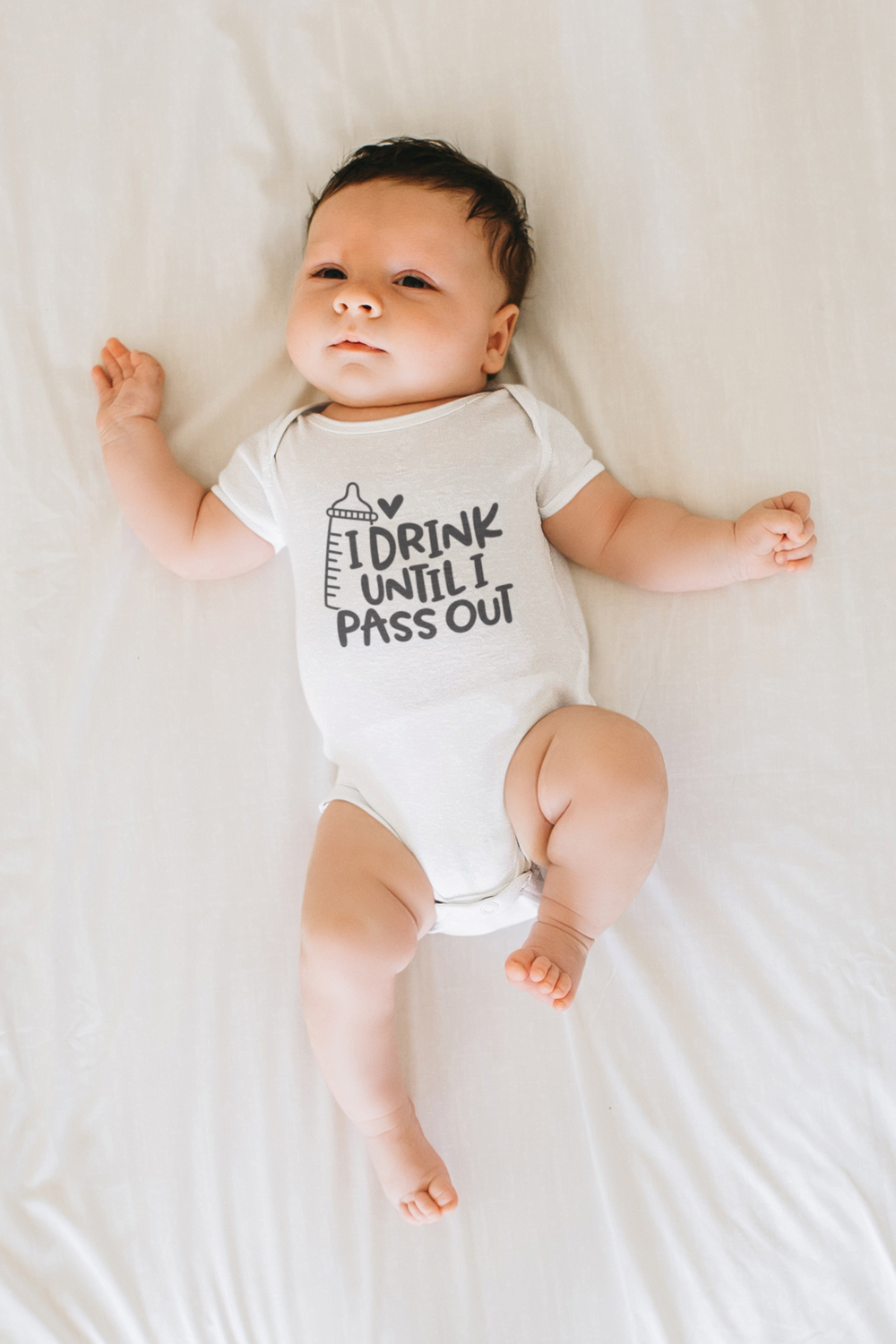 A baby lies on a bed wearing an I Drink Until I Pass Out onesie. Infant fine jersey bodysuit with 100% cotton fabric, ribbed knitting bindings, and plastic snaps for easy changing access. From Worlds Worst Tees.