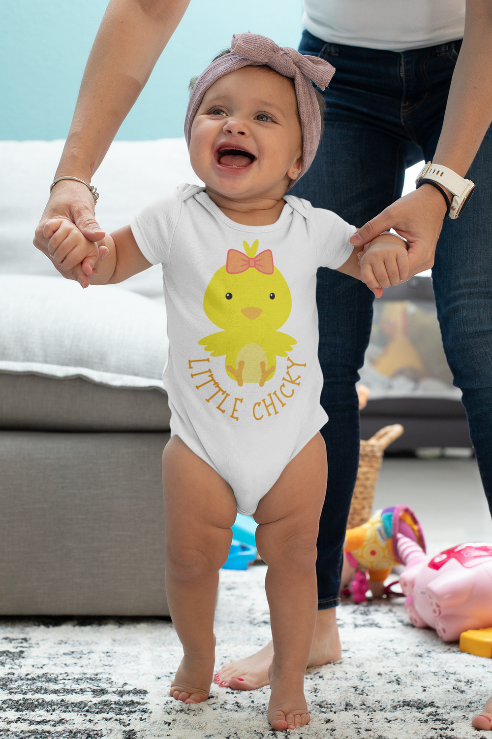 A baby in a Little Chicky Onesie standing in front of a woman's hands, showcasing a soft and comfortable infant bodysuit made of ring-spun cotton for ultimate coziness. Features lap shoulders for easy adjustments.