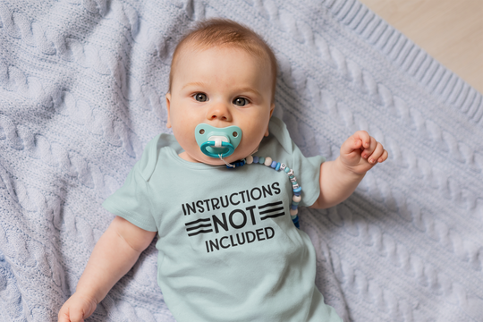 A baby lies on a blanket, wearing an Instructions Not Included Onesie from Worlds Worst Tees. This infant fine jersey bodysuit is 100% cotton, featuring ribbed knitting for durability and plastic snaps for easy changing access.