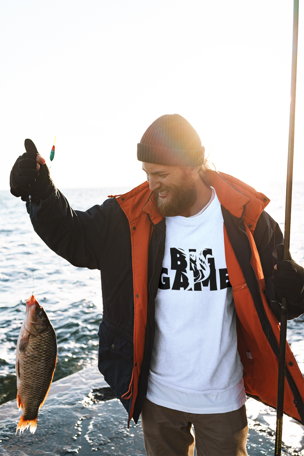 Alt text: BIG GAME FISHER Tee: A man holding a fish and fishing pole by a lake, embodying Comfort Colors' ring-spun cotton tee with a relaxed fit and durable stitching.