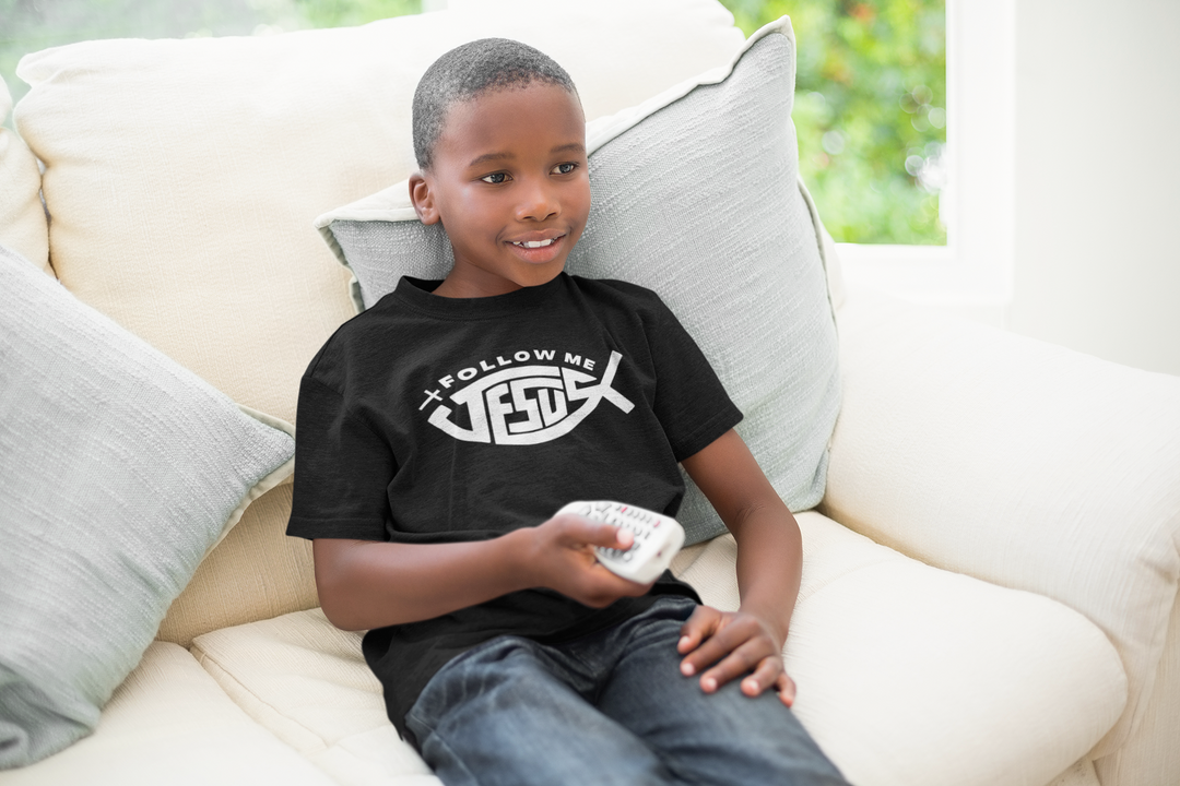 A boy sitting on a couch holding a remote control, wearing a Jesus Follow Me Kids Tee. Comfortable 100% cotton, classic fit, ideal for everyday wear. Visible features: tee, remote control, couch.